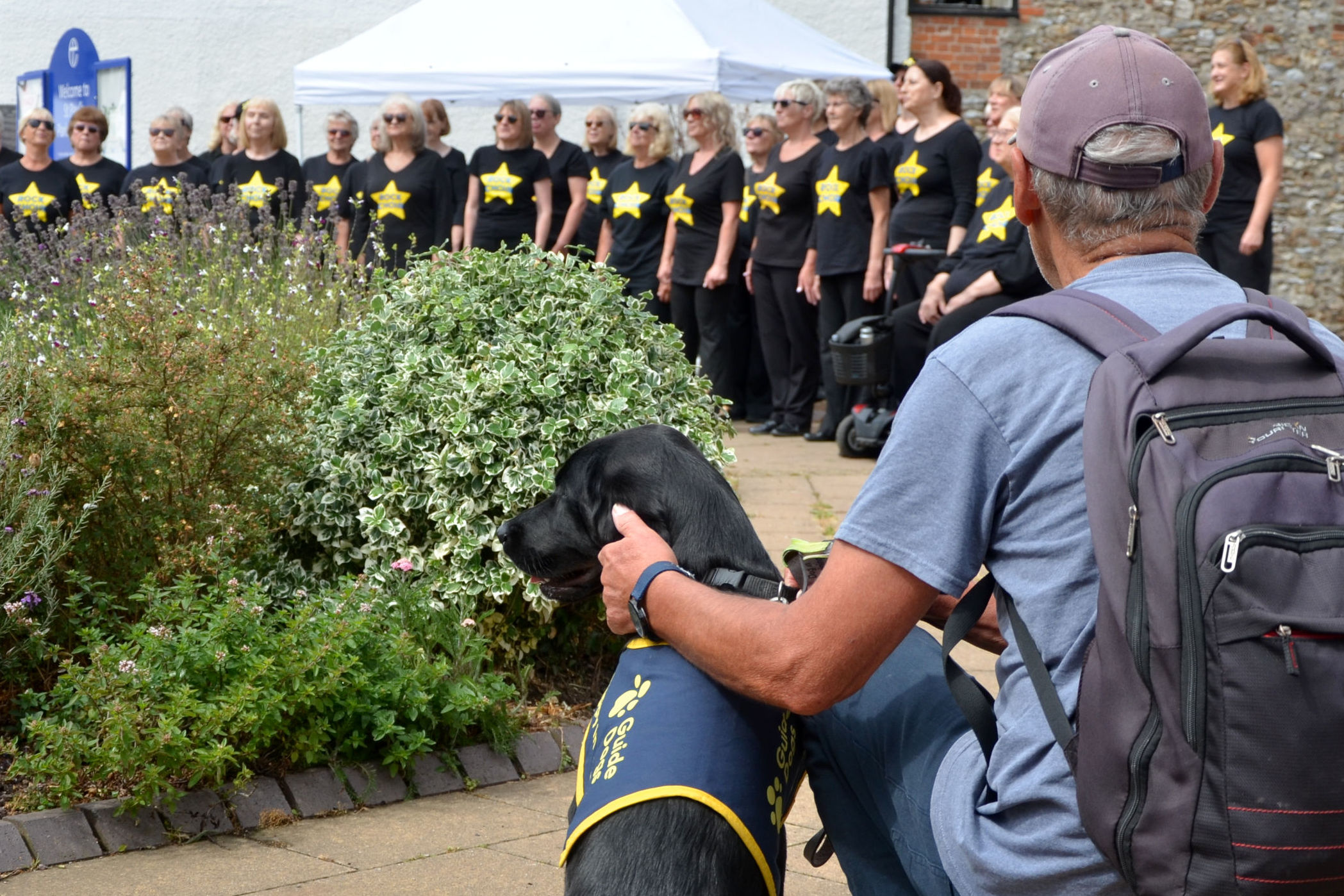 The Rock Choir singing outside St Pauls Church in Honiton watched by man with Guide Dogs for the Blind trainee dog.